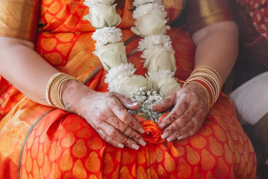 Mains de la mariée ornées de henné et de bracelets dorés sur un sari orange, guirlande de fleurs
