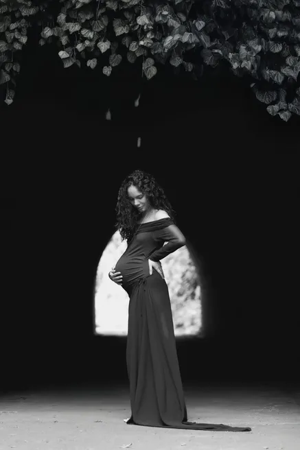 En noir et blanc, future maman en robe longue sombre, en silhouette sous une arche de lumière