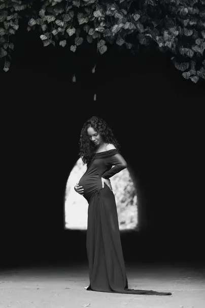 En noir et blanc, future maman en robe longue sombre, en silhouette sous une arche de lumière