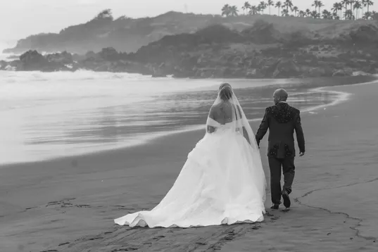 En noir et blanc, les mariés marchent sur le sable, la traîne derrière eux