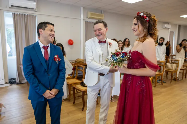 Le marié en costume bleu attend la mariée en robe rouge, accompagnée par un homme en costume blanc
