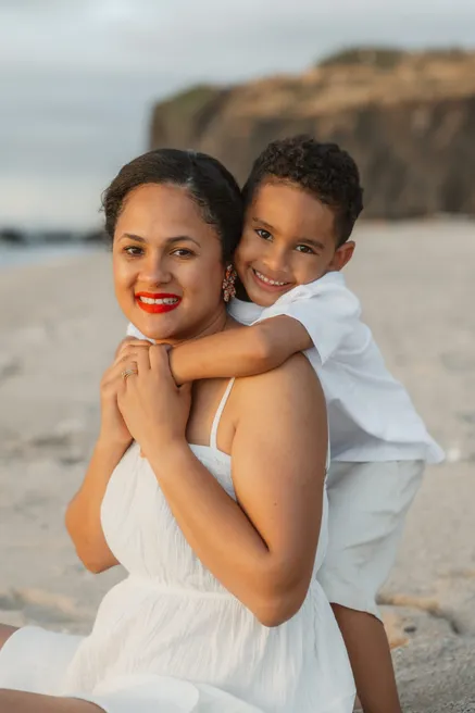 Une mère au rouge à lèvres vif sourit, son fils accroché à ses épaules sur la plage