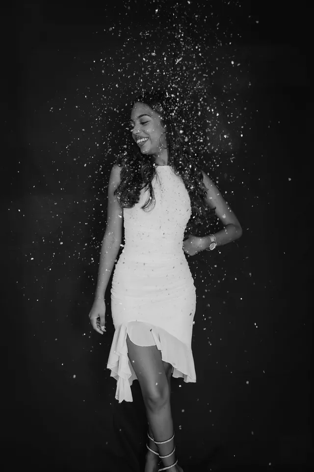 En noir et blanc, une femme en robe blanche rit sous une pluie de confettis, sur fond noir