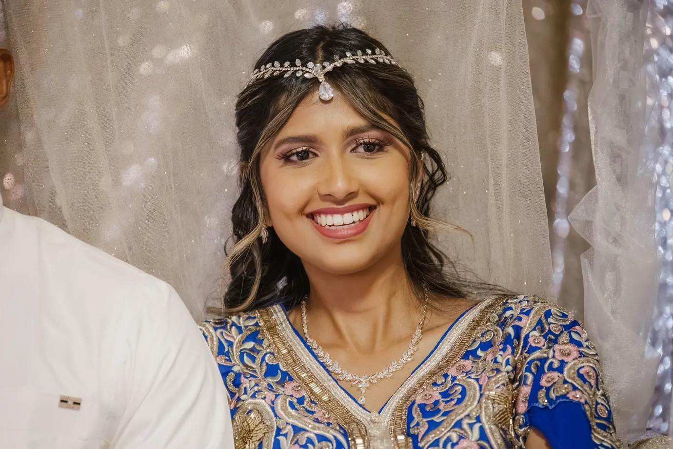 La mariée en tenue bleue brodée et bijou frontal sourit devant un voile pailleté