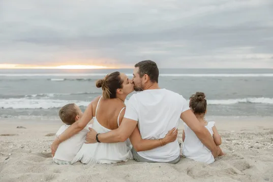 De dos, une famille assise sur le sable face à la mer, les parents s’embrassent