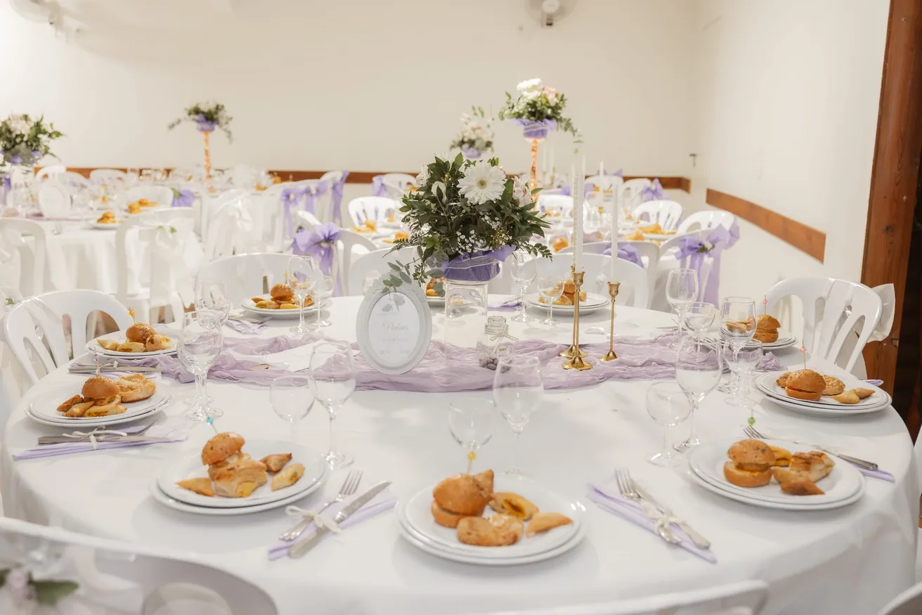 Table ronde dressée de nappes blanches, voilage lilas, verres et petits fours
