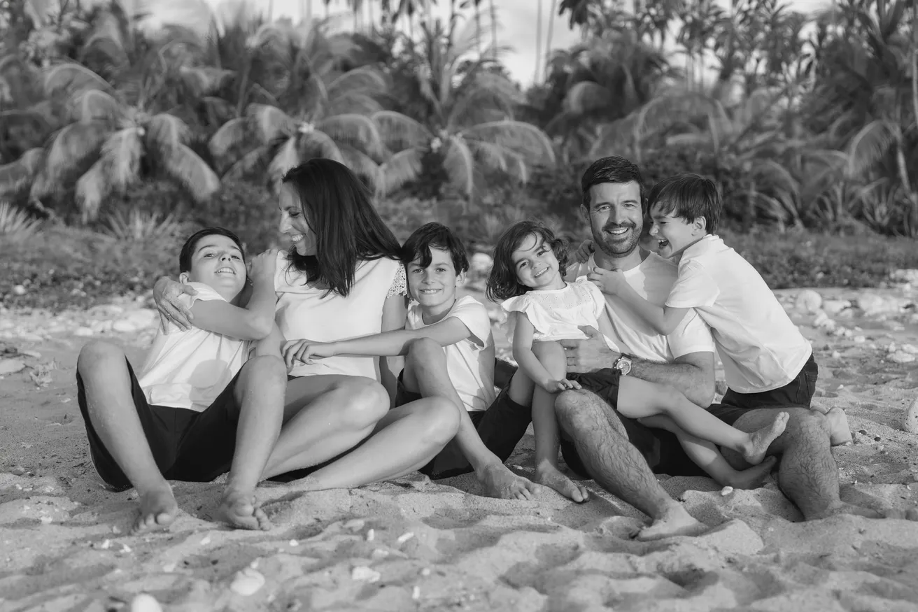 En noir et blanc, une famille de cinq assise sur le sable s’enlace en riant, les palmiers derrière