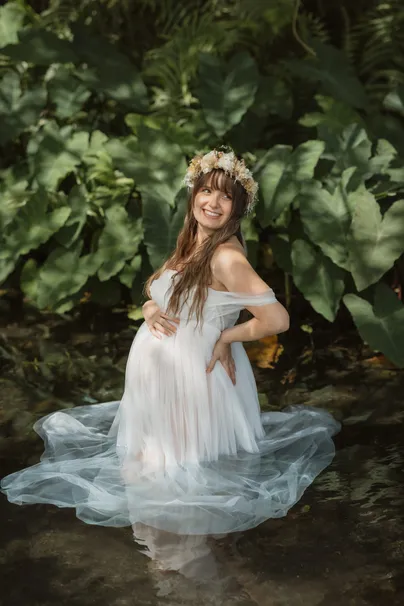 Future maman couronnée de fleurs assise dans l’eau, robe de tulle étalée, feuillage derrière