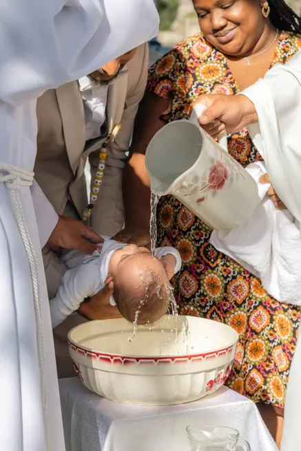 Un bébé penché au-dessus d’une vasque reçoit l’eau versée d’une cruche, sa marraine à côté