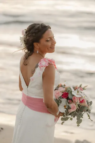 La mariée de dos regarde par-dessus l’épaule, ceinture rose et bouquet, devant la mer