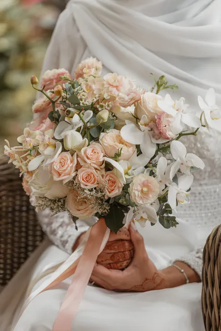 La mariée tient un bouquet de roses pêche et d’orchidées blanches, noué d’un ruban