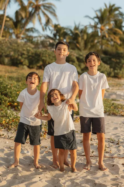 Quatre frères en tee-shirts blancs rient ensemble, pieds nus dans le sable, devant la végétation