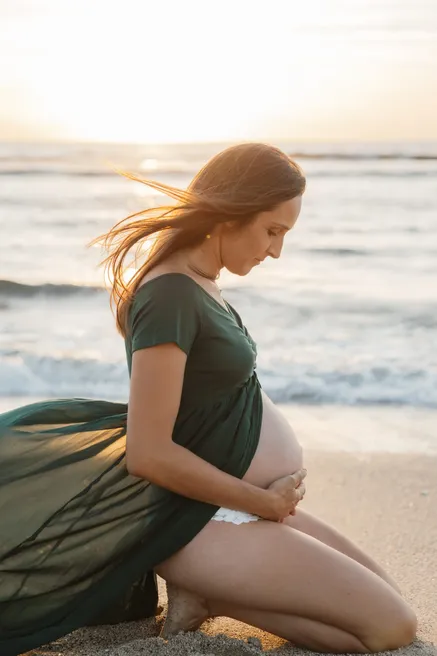Future maman à genoux sur le sable, robe verte, les cheveux au vent devant la mer