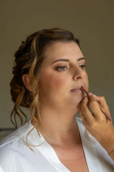 La mariée se fait appliquer un crayon sur les lèvres, cheveux relevés et chemise blanche
