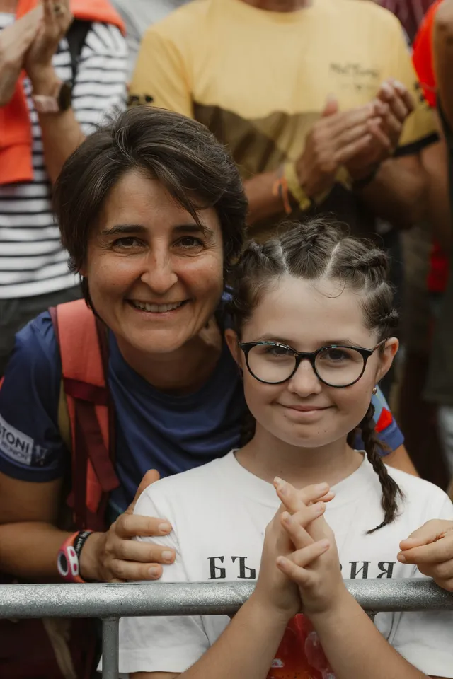 Une maman et sa fille à lunettes, penchées sur la barrière, attendent l’arrivée des coureurs