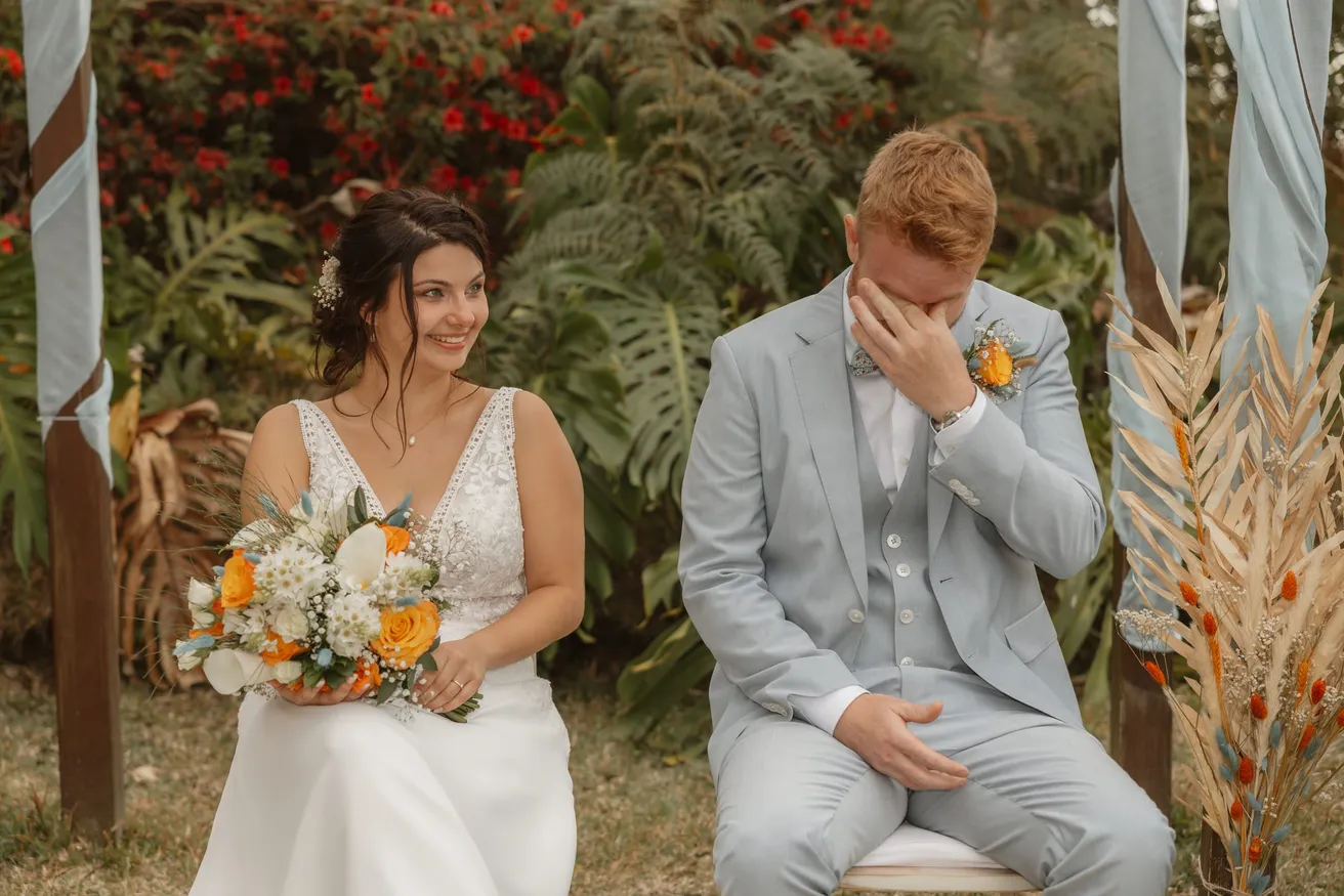 Le marié en costume bleu clair essuie une larme à côté de la mariée souriante