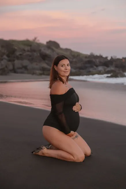 Future maman à genoux sur le sable noir, body noir, ciel rose derrière elle