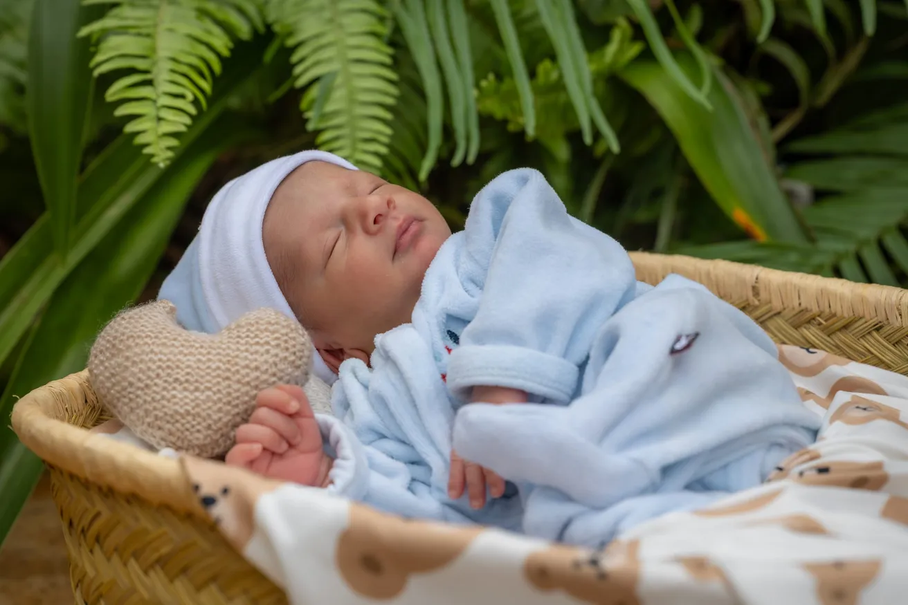 Le nouveau-né en bonnet et pyjama bleus dort dans un panier, près d’un cœur tricoté, sous les fougères
