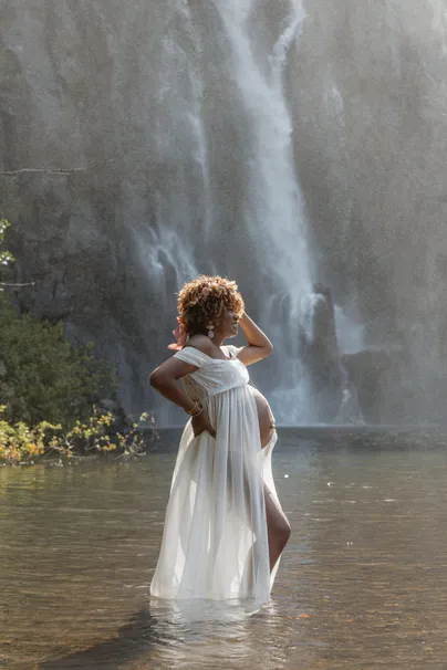 Future maman en robe blanche debout dans l’eau, main dans les cheveux, cascade derrière elle