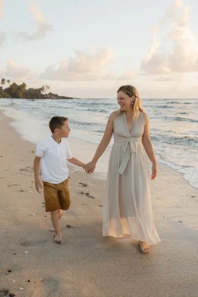 Une mère en longue robe beige marche main dans la main avec son fils sur le sable