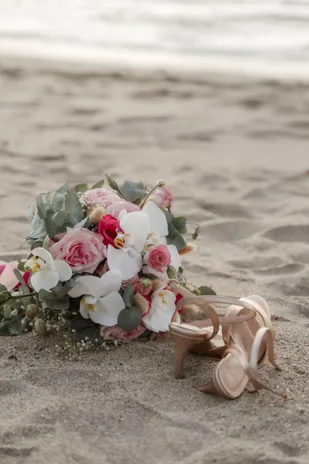 Bouquet de roses et d’orchidées posé sur le sable, à côté d’une paire d’escarpins nude
