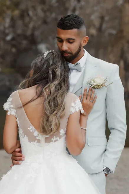 Le marié en costume gris clair enlace la mariée vue de dos, robe au dos de dentelle