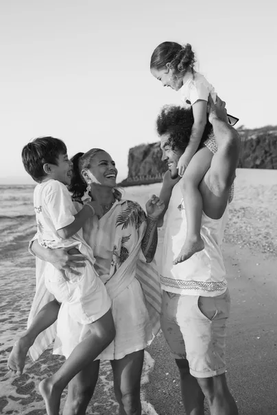 En noir et blanc, un père porte sa fille sur les épaules et la famille rit au bord des vagues