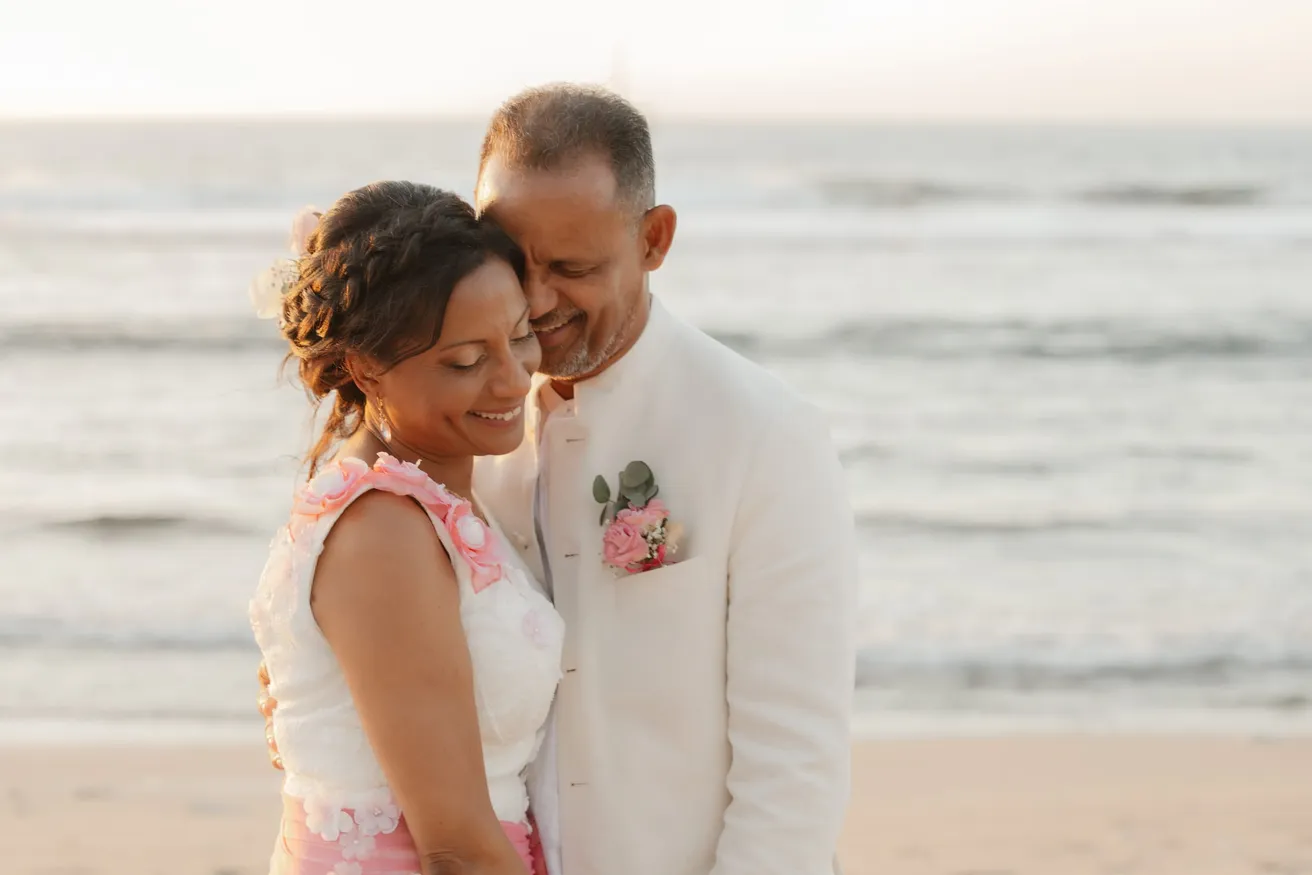 La mariée pose la tête contre le marié en veste blanche, face à l’océan au couchant