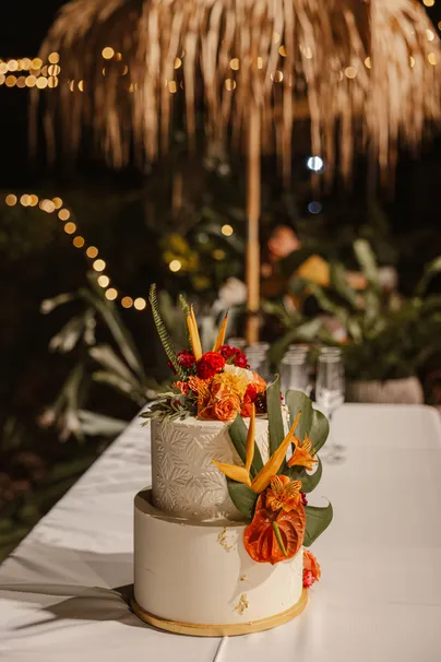Gâteau de mariage à deux étages décoré de fleurs tropicales orange, sous une paillote illuminée