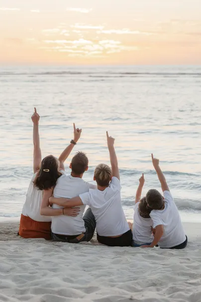 Une famille assise de dos sur le sable lève le bras vers le soleil couchant