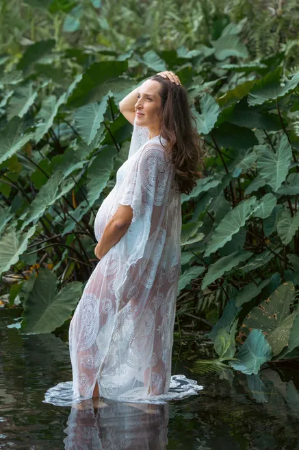 Future maman en robe transparente à fleurs debout dans l’eau, entre de grandes feuilles vertes