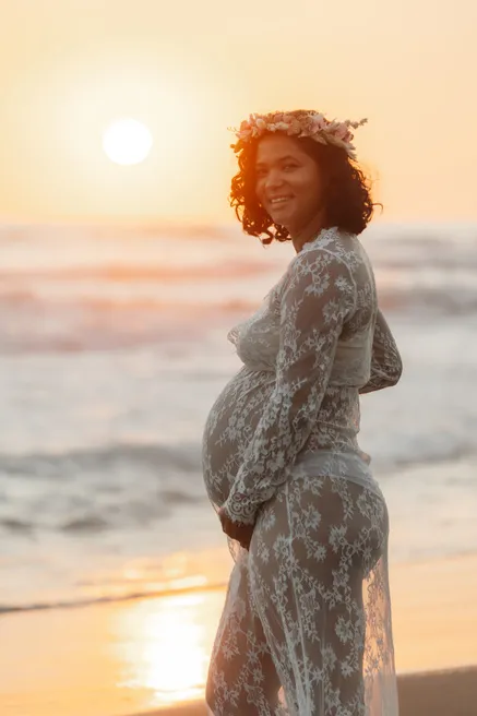 Future maman en dentelle et couronne de fleurs, tenant son ventre, soleil couchant derrière