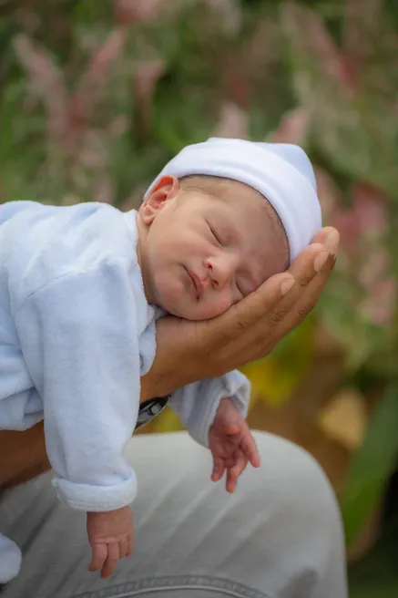 Le nouveau-né en bonnet blanc dort à plat ventre sur la main d’un adulte, devant la verdure