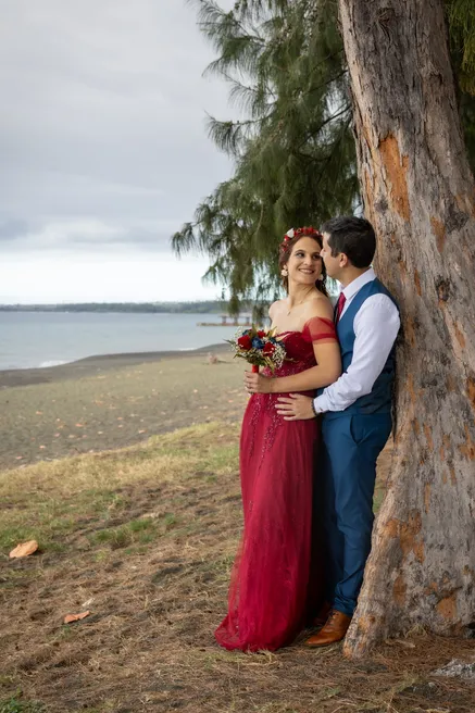 Les mariés enlacés contre un tronc d’arbre au bord d’une plage de sable noir, la mariée en rouge