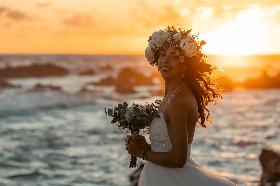 La mariée en couronne de fleurs rit face à la mer, au soleil couchant