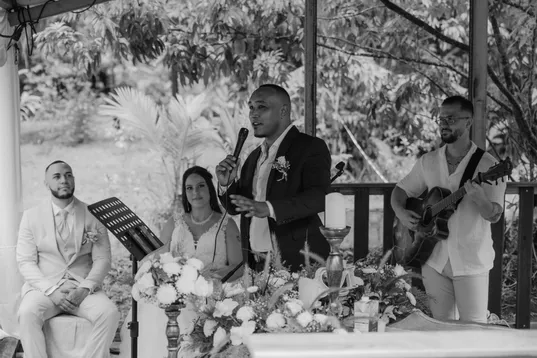 En noir et blanc, un invité chante au micro près des mariés assis, accompagné d’un guitariste