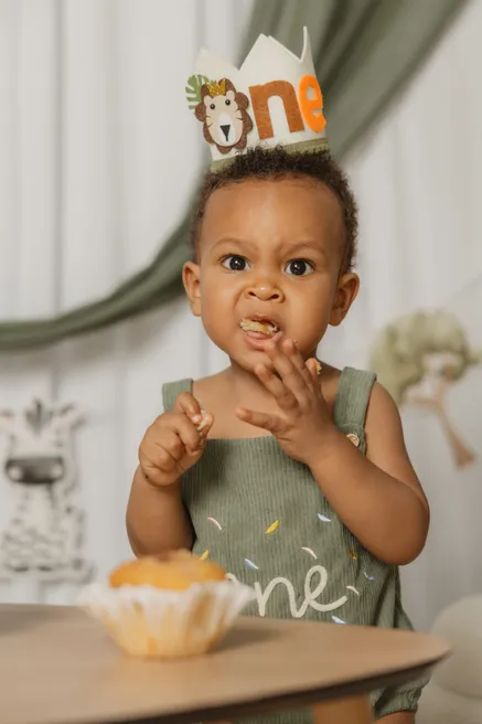 Un petit garçon coiffé d’une couronne « one » goûte son muffin, les doigts à la bouche