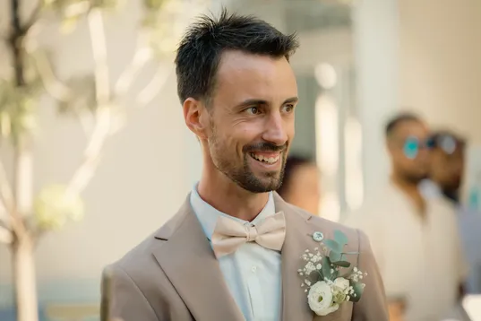 Le marié en costume beige et nœud papillon clair sourit, boutonnière fleurie