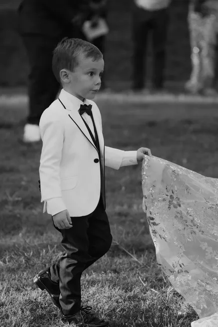 En noir et blanc, un petit garçon en smoking blanc porte la traîne de la robe de la mariée