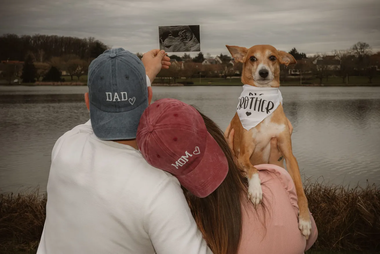 Un couple de dos en casquettes DAD et MOM brandit une échographie, leur chien au bandana