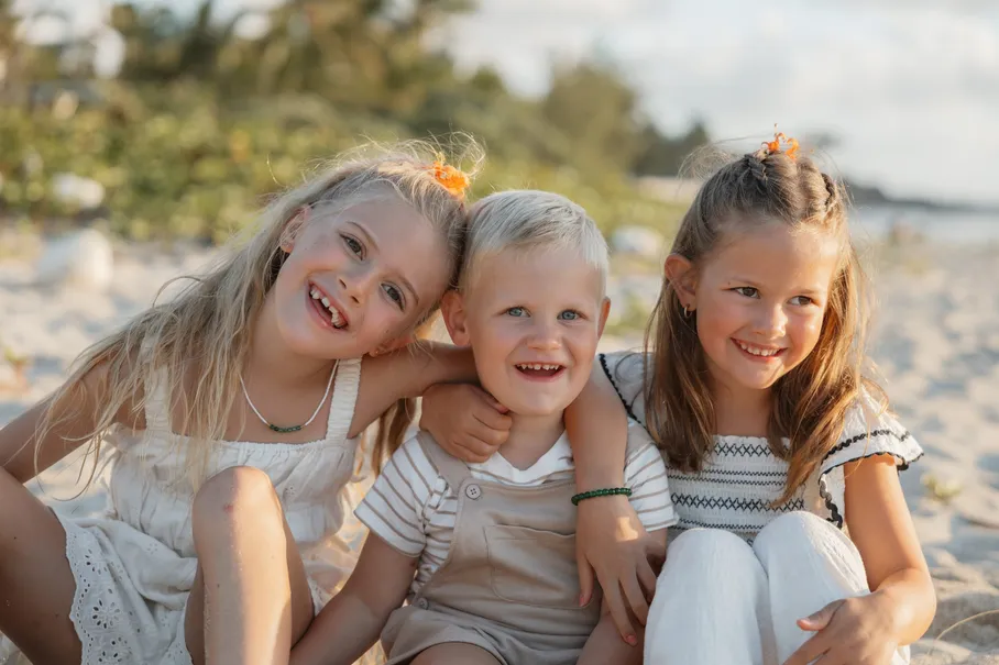 Trois enfants blonds serrés les uns contre les autres rient, assis sur le sable
