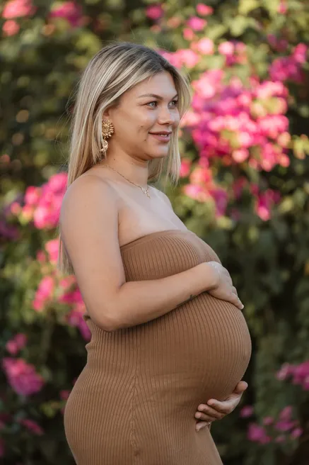 Future maman en robe beige moulante, mains sur son ventre, devant des bougainvilliers roses