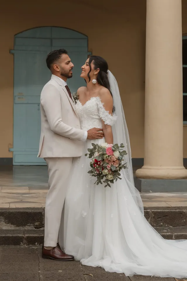 Les mariés se regardent sous les colonnes d’une maison créole aux volets bleus, le bouquet à la main