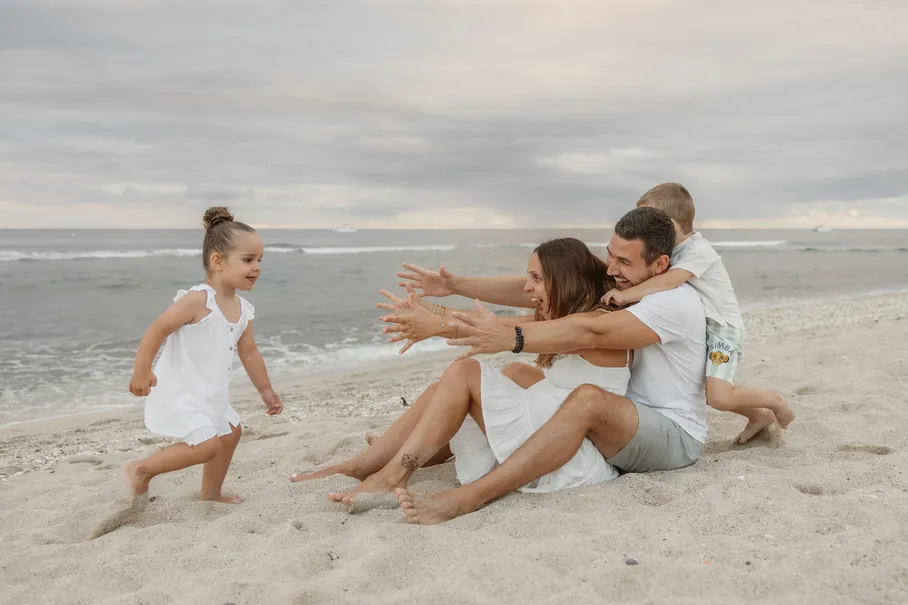 Une petite fille court bras tendus vers ses parents assis sur le sable, un garçon sur le dos du père