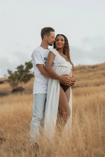 Future maman en robe blanche fendue enlacée par son compagnon, au milieu des herbes sèches