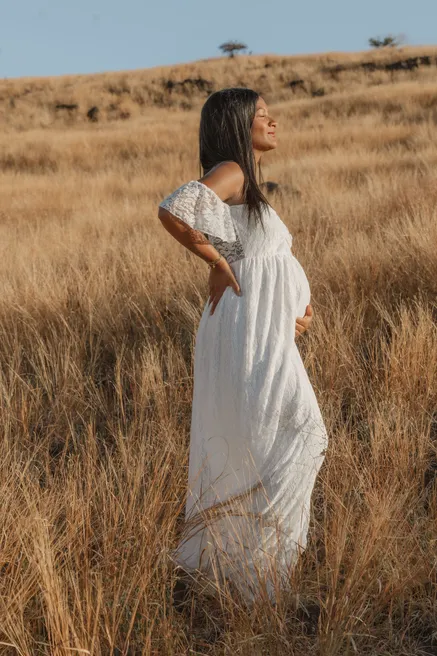 Future maman debout dans les herbes hautes dorées, robe blanche, le visage tourné vers le soleil