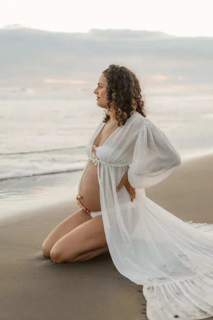 Future maman en robe blanche vaporeuse agenouillée sur le sable, de profil face à la mer