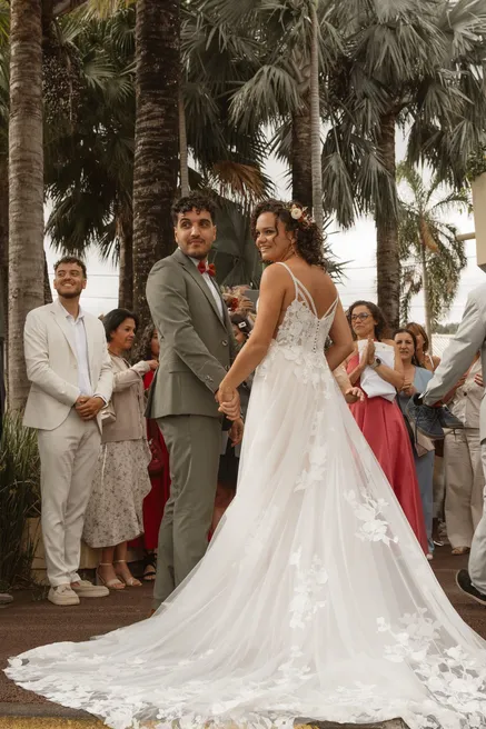 Les mariés face à face devant leurs invités, sous les palmiers, traîne de dentelle étalée au sol