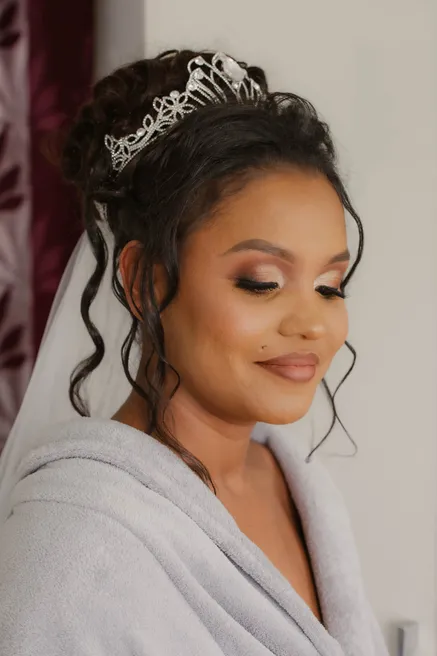 La mariée en peignoir, diadème et voile dans les cheveux, sourit les yeux baissés