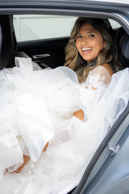 La mariée rit assise dans une voiture, sa robe de tulle débordant de la portière